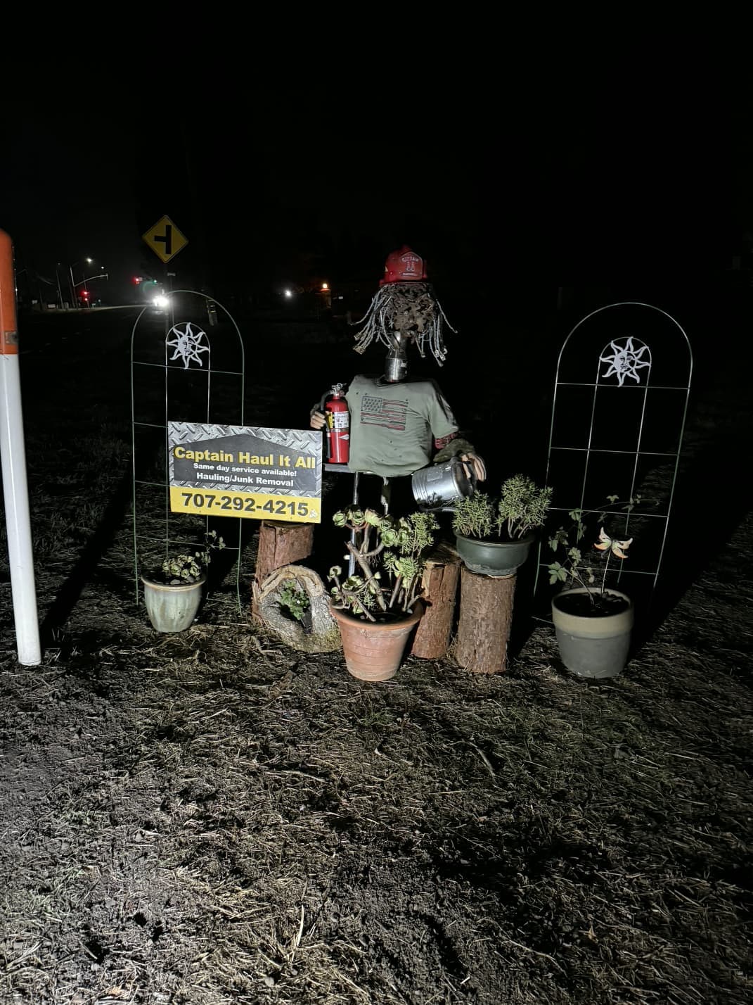Nighttime outdoor scarecrow display with potted plants and decorative lighting.
